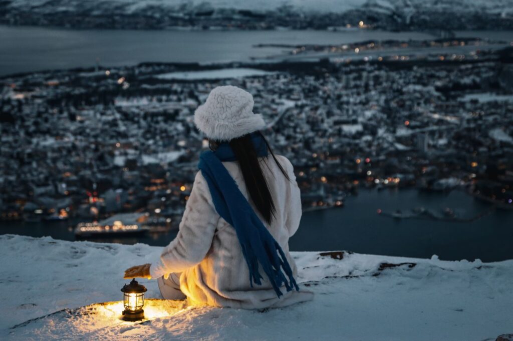 A person sitting on a snowy cliff with a lantern, overlooking Tromsø, Norway at night.